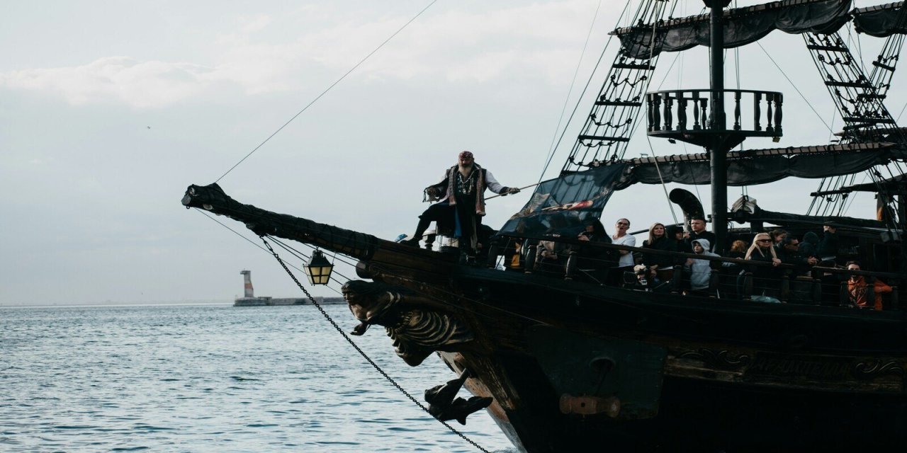Darj pirate ship with cannon at the front deck. Man standing on the edge. Grey skies and blue sea with rippled waves. 
